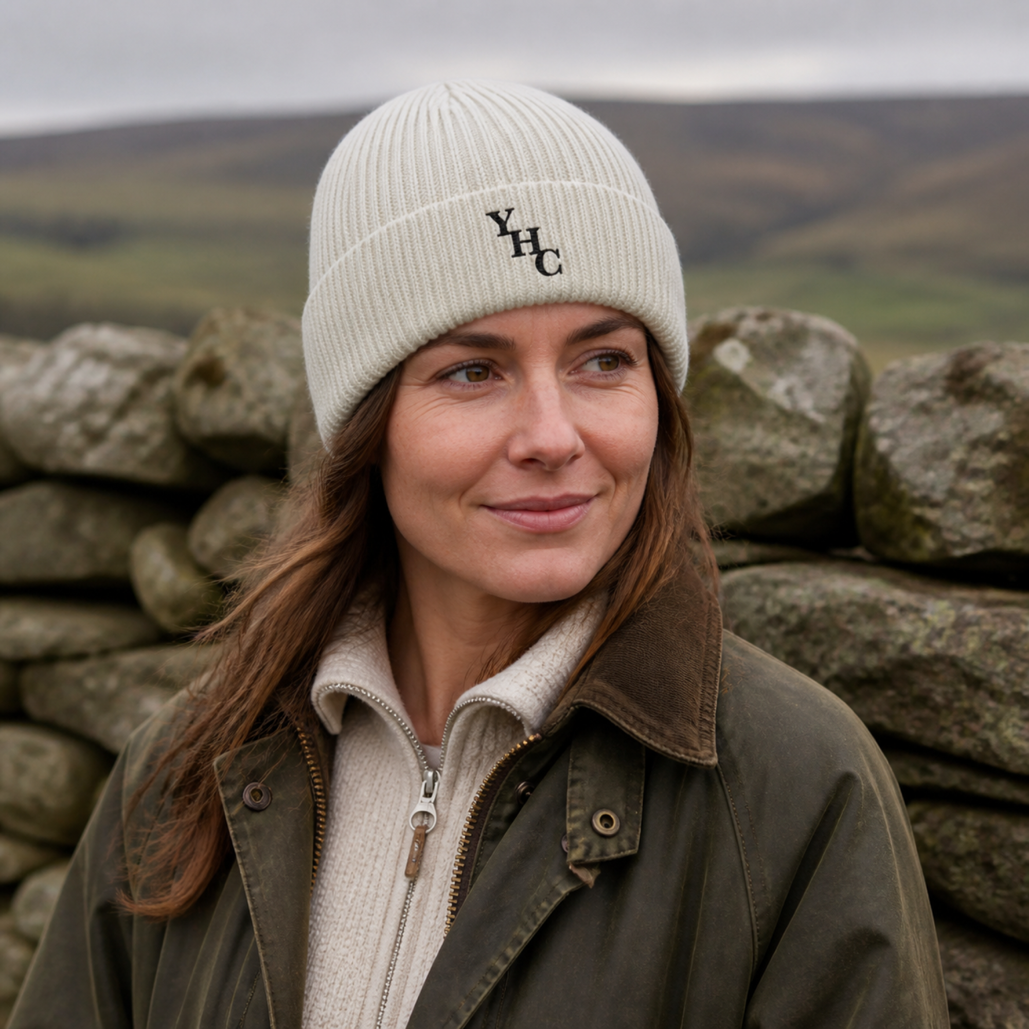 Raw Yorkshire Heart Clothing ribbed beanie with true-black embroidered YHC monogram worn beside a Yorkshire dry-stone wall